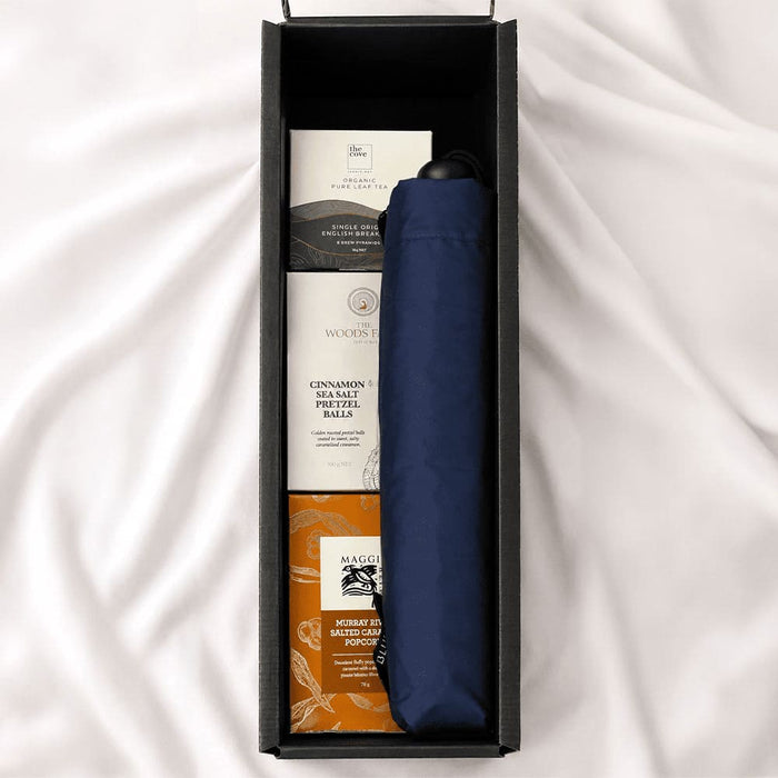 The Blunt Umbrella All Weather Set - A navy umbrella is nestled in a box with treats like cinnamon balls and other snacks. The box has a sleek design and logo.
