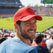 Classic 5 Panel Trucker Mesh Cap - A man wearing a red Classic 5 Panel Trucker Mesh Cap smiles at a sports event, with a crowd in the background.