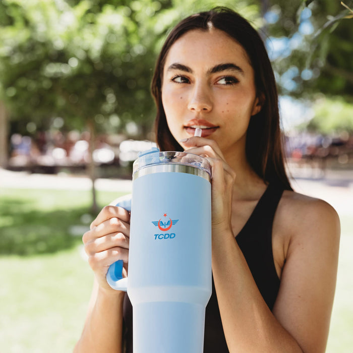 The Hippo 1.2L Mug - A drink bottle in light blue with a handle, featuring a logo and a clear lid, held by a person outdoors.