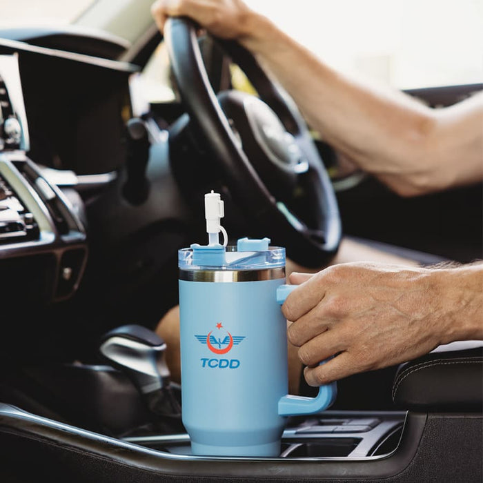 The Hippo 1.2L Mug - A drink bottle in light blue, featuring a handle and a straw, positioned in a car's centre console.