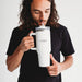 The Hippo 1.2L Mug - A drink bottle in white with a transparent lid and straw, featuring a logo, held by a person.