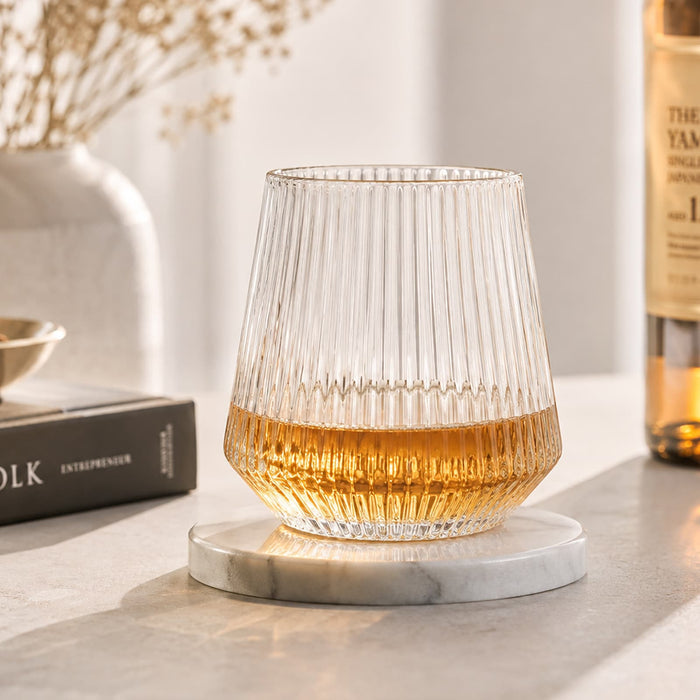 Velora Tumbler by The Line Edit. - A drink bottle with a ribbed clear glass design sits on a marble coaster, featuring a golden liquid inside.
