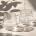 Velora Tumbler Gift Set by The Line Edit. - A drink bottle featuring a ribbed design, made of clear glass, with water being poured into two glasses.