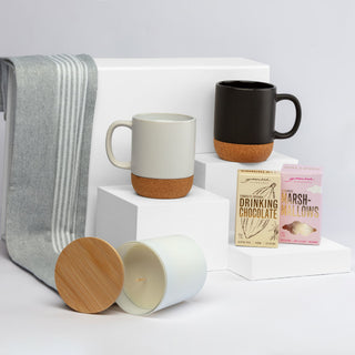 The Warm Welcome - A collection of two mugs in grey and black with cork bases, a candle, and chocolate and marshmallow packets on white display.