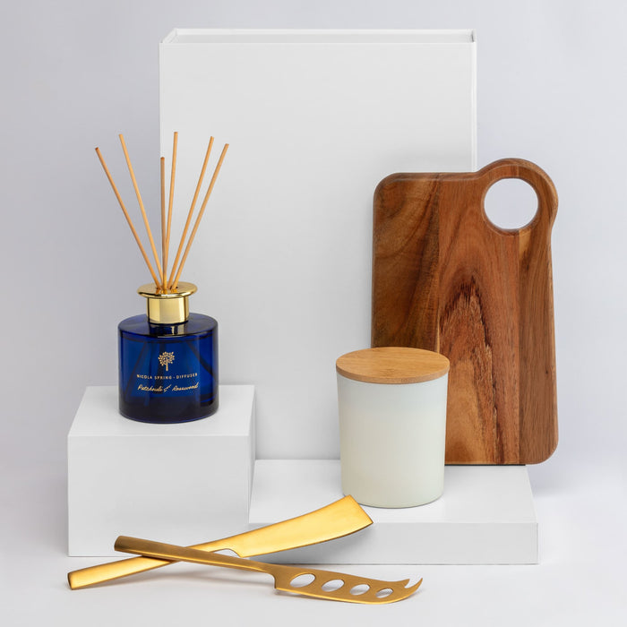 The Host Edit - A collection of homeware items including a blue diffuser, wooden board, gold utensils, and a white candle jar on white pedestals.