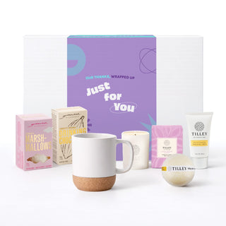 The Cosy Indulgence - A gift set containing a white mug with a cork base, marshmallow treat, and various skincare products in pastel packaging.