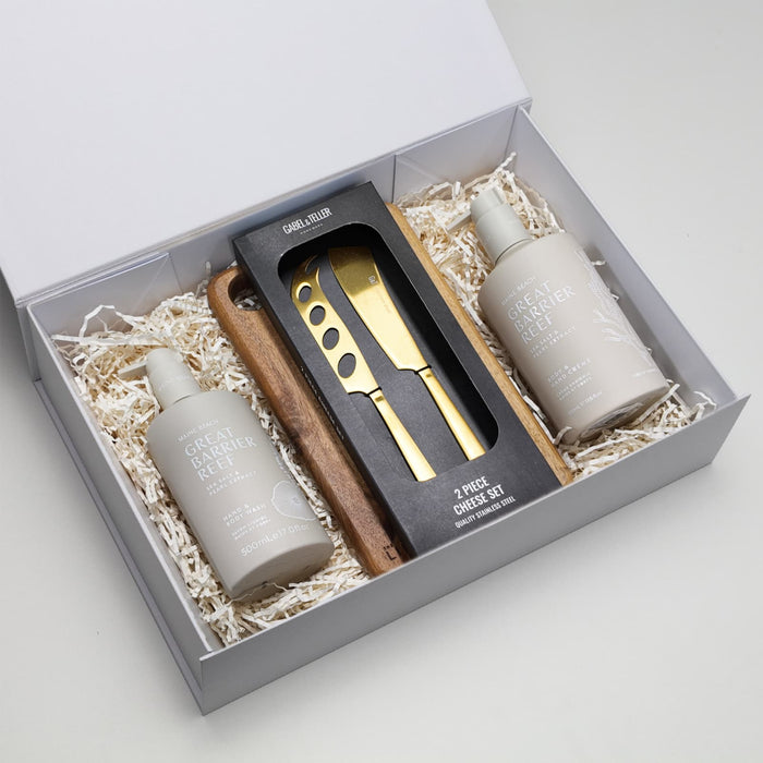 The Pamper & Entertain Edit - A gift box containing two cream bottles and a wooden cheese knife set, arranged on shredded paper.