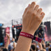 Event Wrist Band - A hand wearing a vibrant pink and purple wristband, with a blurred concert crowd in the background and a stage visible.