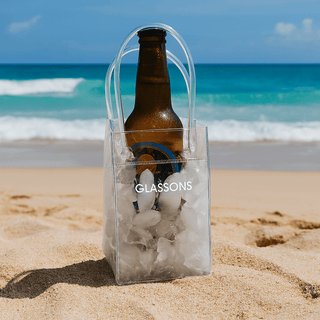 Icy Bags - Small - A clear cooler bag filled with ice, holding a brown drink bottle, placed on sandy beach by the ocean. It has a logo.