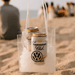 Icy Bags - Large - A clear cooler bag filled with ice and two cans, featuring a logo. Set on a sandy beach with people in the background.