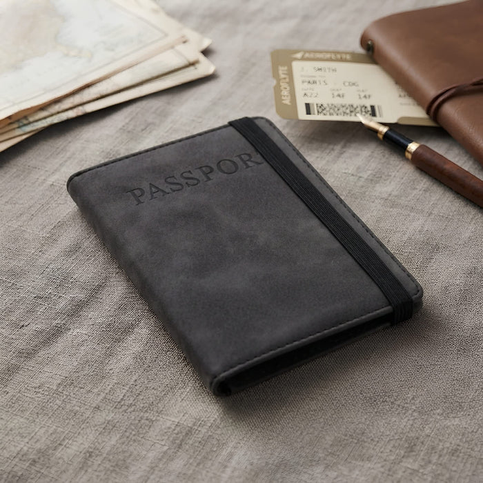 Traveller Passport Wallet - A black passport wallet with an elastic band, resting on a textured surface alongside travel documents and a pen.