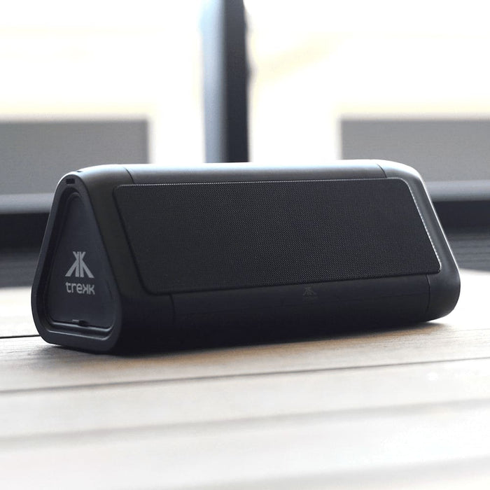 Trekk Outdoor Speaker & Wireless Powerbank - A black outdoor speaker with a triangular shape and a logo, resting on a wooden surface.