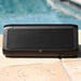 Trekk Outdoor Speaker & Wireless Powerbank - A black outdoor speaker with a mesh front, placed on a stone surface by a pool. It has branding on the side.