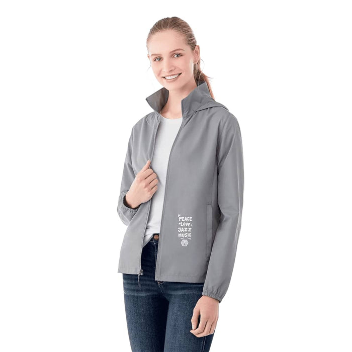 Women's TOBA Packable Jacket - A woman wearing a grey packable rain jacket with a hood, featuring a logo and text on the front.