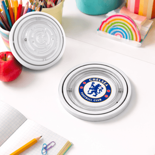 The Amazing Fidget - A circular coaster featuring a logo on a table, surrounded by stationery, an apple, and colourful paper.